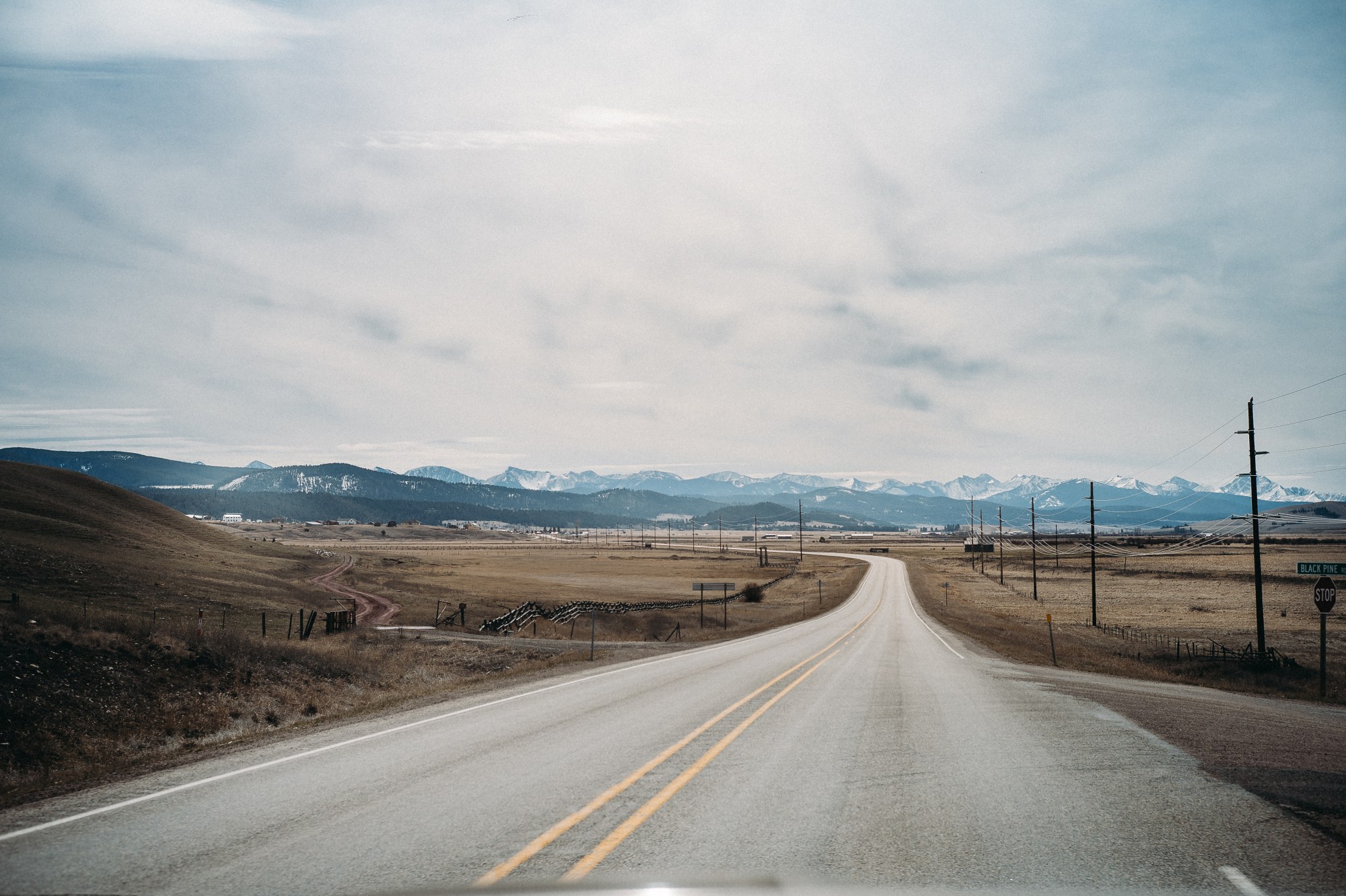 A straight rural highway stretches toward snow-capped mountains in the distance, with dry grassland and utility poles flanking both sides under a cloudy sky.