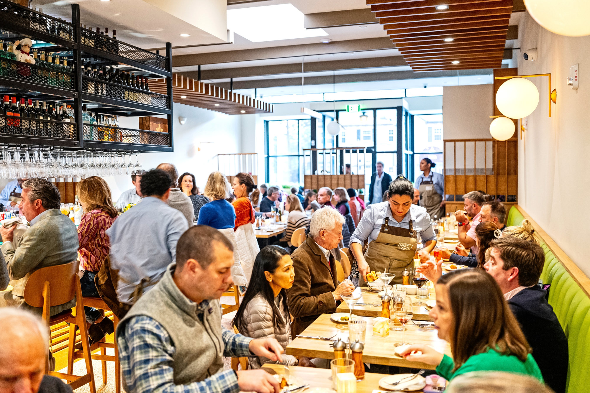 A busy restaurant dining room filled with guests seated at long communal tables being served by staff members, with modern wood and glass architectural features and large windows visible in the background.