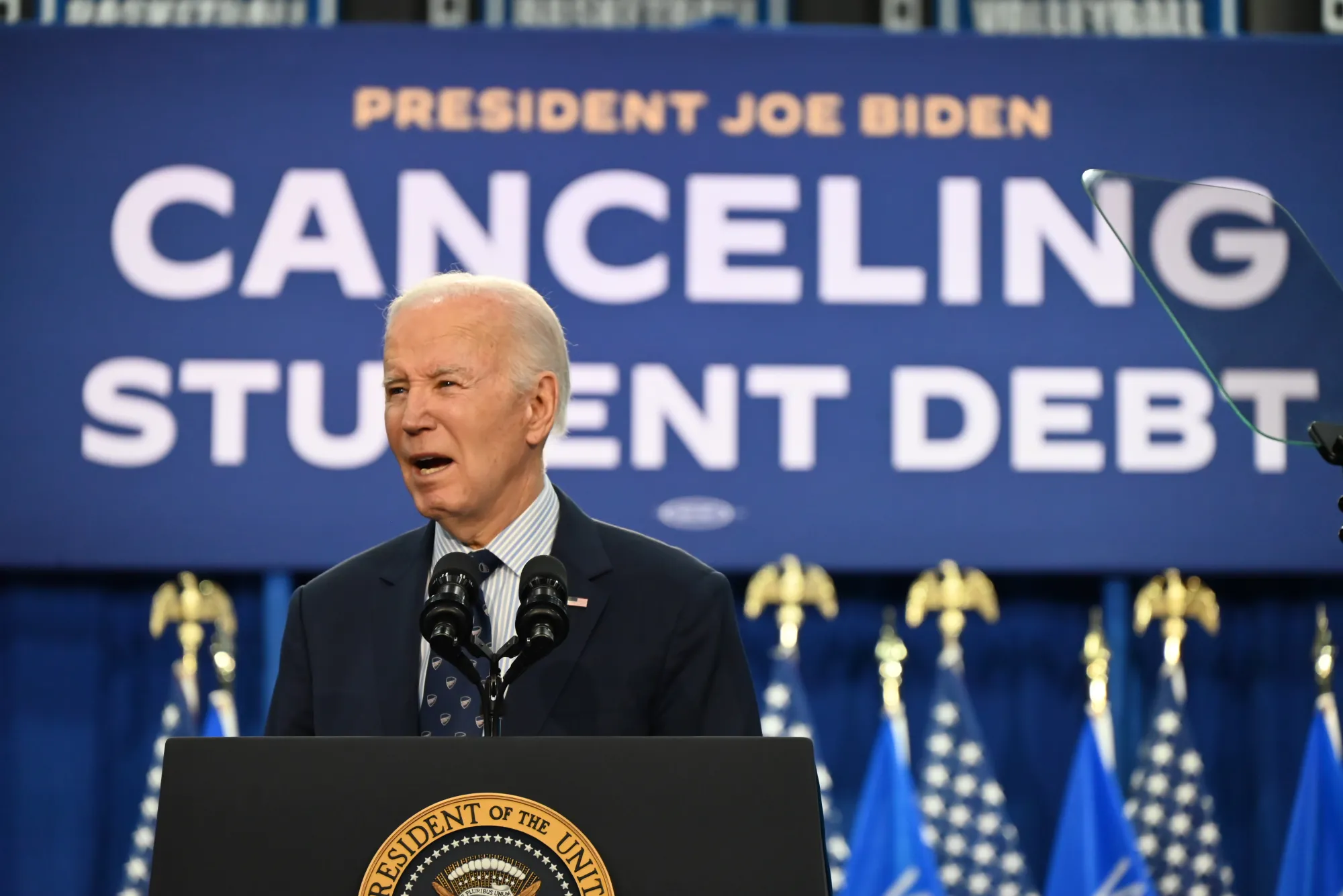President Joe Biden speaks at a podium with the presidential seal during an announcement about canceling student debt, with a blue backdrop and American flags visible in the background.